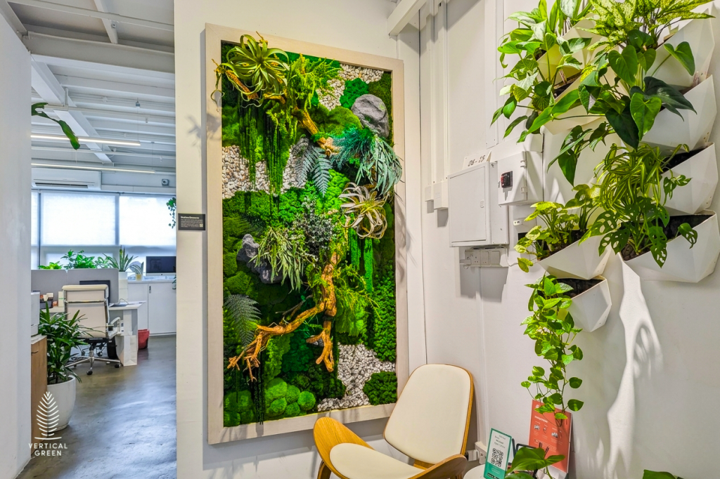 Creative office interior showcasing a framed preserved moss wall art piece with wood accents and pebbles by Vertical Green Malaysia. A vertical wall planter system with cascading green foliage hangs adjacent to a minimalist lounge chair.